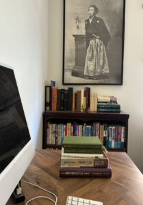 A stack of Japanese-language sources used in researching The Last Shogun sits on a writing desk beneath a portrait of Sakamoto Ryōma, with a shelf of Shinsengumi and Bakumatsu reference works in the background.